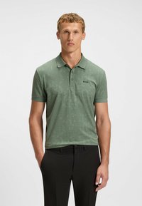 Grön polo t-shirt med ett texturerat geometriskt mönster. Har krage och korta ärmar, kombineras med svarta byxor.
