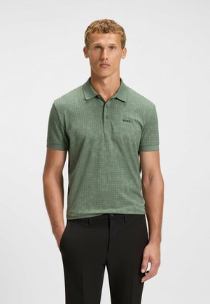 Grønt polo-shirt med et struktureret geometrisk mønster. Har en kravehals og korte ærmer, kombineret med sorte bukser.