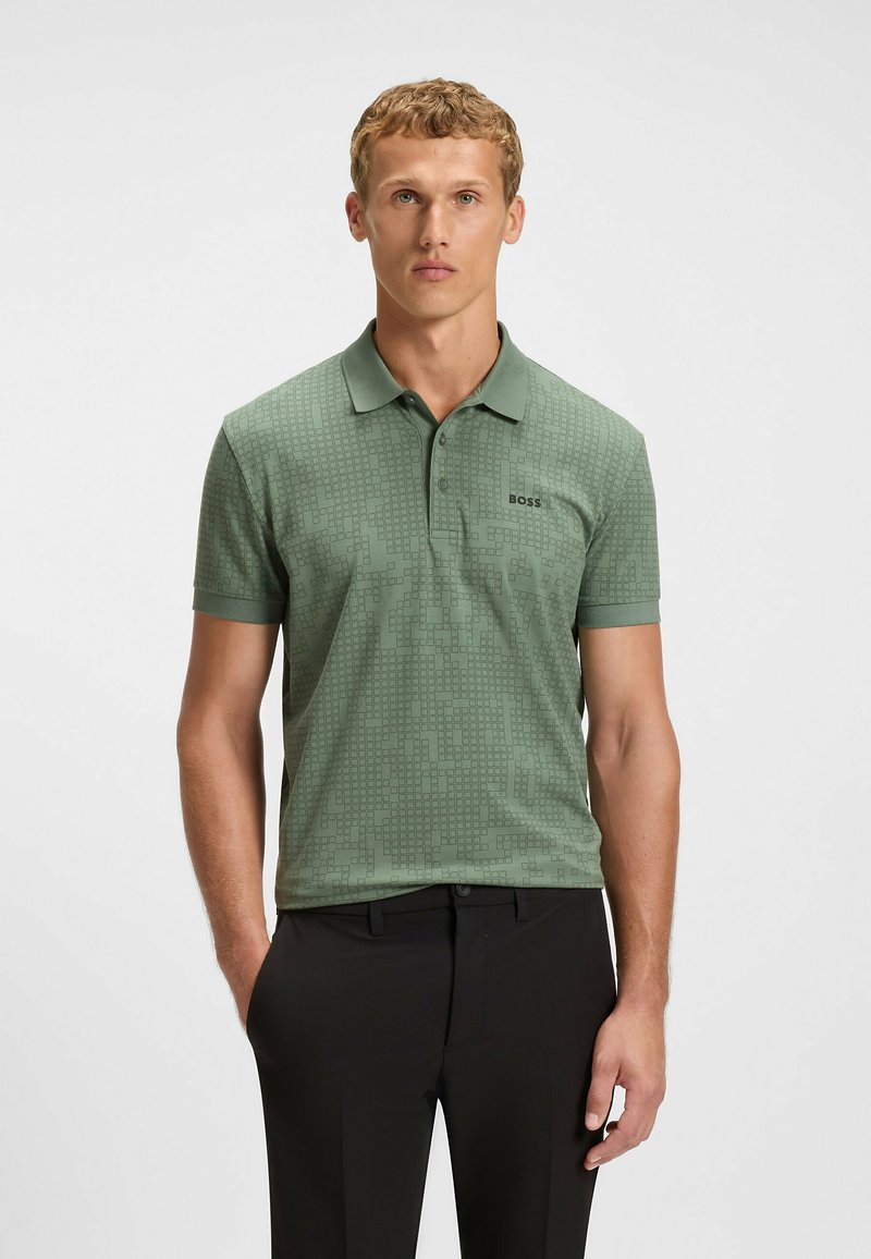 Grön polo t-shirt med ett texturerat geometriskt mönster. Har krage och korta ärmar, kombineras med svarta byxor.