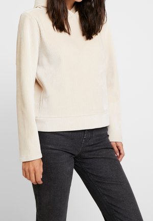 Woman wearing a cream ribbed hoodie and black jeans standing against a plain white background, shown from shoulders to hips.