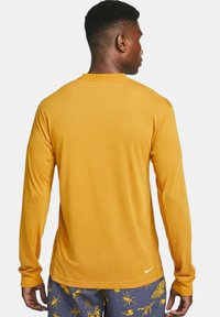 Long-sleeve, mustard-yellow Nike shirt with a rounded neckline, smooth texture, and minimalistic design, featuring a small logo on the back.