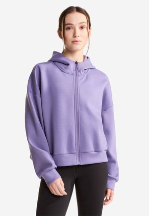 Ung kvinde iført en lavendelfarvet hættetrøje med lynlås, med hætten nede, og sorte leggings, står foran en hvid baggrund.