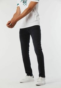 Jean en denim foncé ajusté associés à un t-shirt blanc avec des accents verts, portés avec des baskets blanches, mettant en avant un style décontracté.