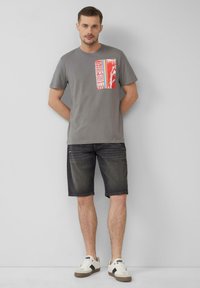 Grijze katoenen T-shirt met een rode grafische print, gecombineerd met donkere denim shorts. Witte sneakers met zwarte accenten maken de outfit compleet.