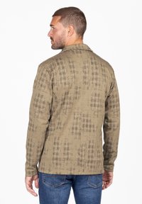 Chaqueta de cuadros marrón con tejido texturizado, mangas largas y cuello. Llevada sobre unos vaqueros azules, mostrando un sutil patrón de espiga.