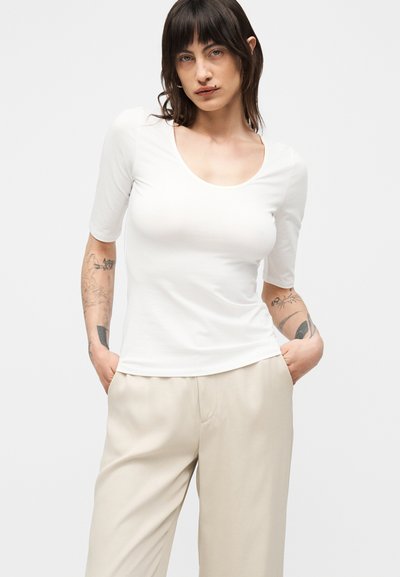 Mujer con cabello oscuro y tatuajes que lleva una camiseta blanca de manga corta con escote redondo y pantalones anchos beige con las manos en los bolsillos.