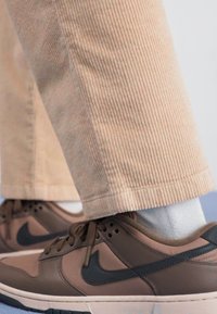 Hnedé tenisky Nike s textúrovaným zvrškom, čiernym logom swoosh a gumovou podrážkou, nosené s svetlo béžovými kordúrovými nohavicami a bielymi ponožkami.