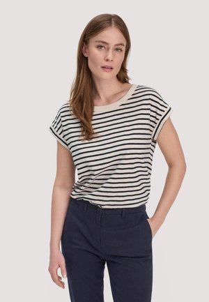 Vrouw die een wit T-shirt met korte mouwen draagt met zwarte horizontale strepen en donkergroene broek, staand met één hand in de zak.