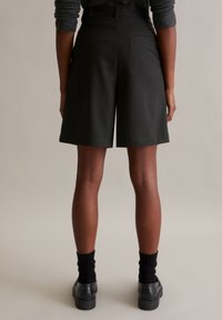 Zwarte op maat gemaakte shorts met een soepele textuur en subtiele streepdessin, met een middelhoge taille en een achterzak, gecombineerd met zwarte sokken en schoenen.