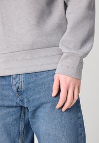Sweatshirt gris clair texturé avec des poignets et un ourlet côtelés, associé à un jean en denim bleu. Gros plan sur une main reposant sur le jean.