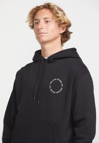 Un sweat à capuche noir en coton, avec une poche avant, un cordon de serrage à la capuche, et un logo rond blanc sur la zone de la poitrine.
