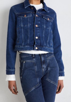 Persona que lleva una chaqueta vaquera azul oscuro ajustada con botones de cobre y vaqueros de talle alto a juego, camisa blanca visible en los puños y la cintura.