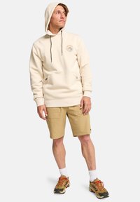 Sudadera crema con cordones, bolsillo frontal y logotipo; combinada con pantalones cortos color crema. Estilizada con zapatillas gruesas y calcetines blancos hasta la mitad de la pierna.