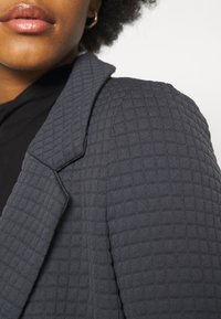 Blazer matelassé gris foncé avec un col structuré et un tissu texturé, présentant des coutures détaillées et une finition lisse.