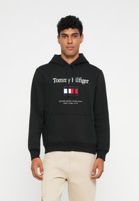 Schwarzer Kapuzenpullover mit weißem "Tommy Hilfiger"-Logo und einem rechteckigen Logo in Rot, Weiß und Blau. Kängurutasche vorne.