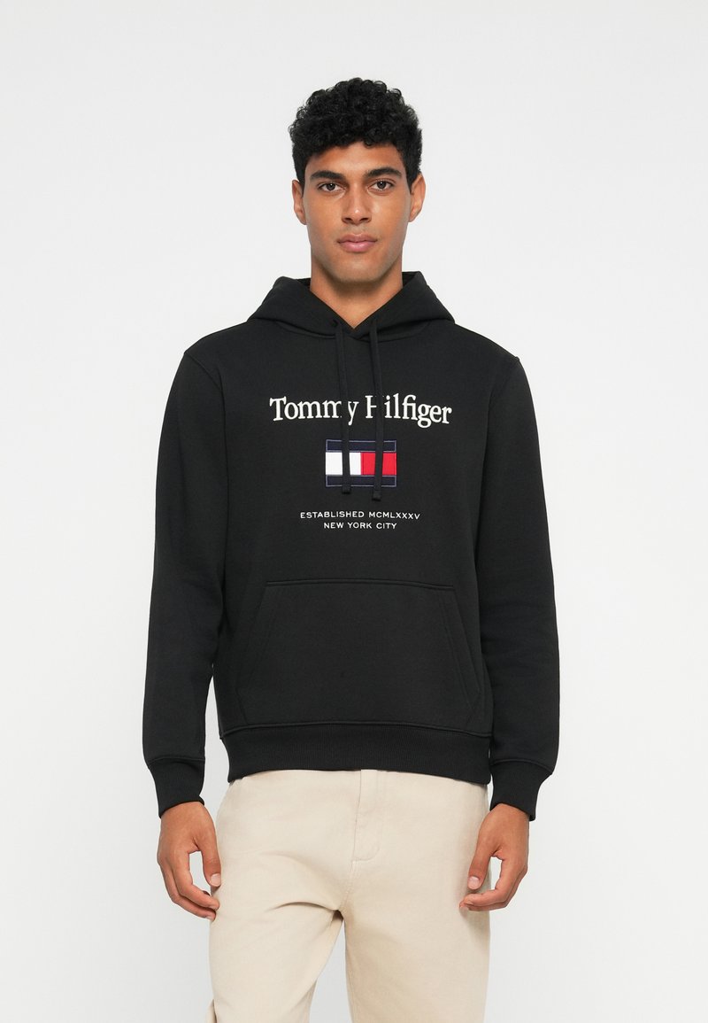 Schwarzer Kapuzenpullover mit weißem "Tommy Hilfiger"-Logo und einem rechteckigen Logo in Rot, Weiß und Blau. Kängurutasche vorne.