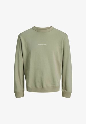 Adolescent aux cheveux bouclés portant une veste à carreaux noire, un sweat-shirt vert, un jean gris ample et des chaussures marron, debout et souriant.