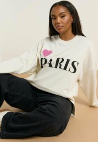 Sudadera de manga larga crema con el texto "I ♥ PARIS" en negro y rosa, combinada con pantalones negros holgados y zapatos blancos. Textura suave.