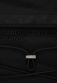 Svart tygväska med präglad text "KARL LAGERFELD JEANS", med en silverklämma och flätade snören för stängning. Slät textur.