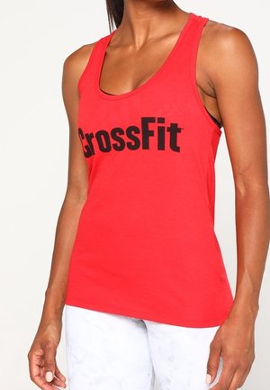 Woman wearing a red sleeveless tank top with "CrossFit" in black text, paired with white pants against a plain background.