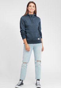 Felpa navy con tasca a marsupio, cappuccio con cordino e polsini a costine, abbinata a jeans chiari strappati e sneakers nere alte.
