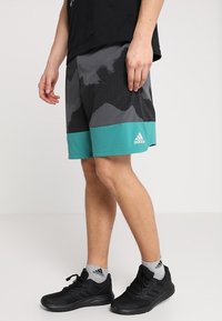Šedé kraťasy Adidas s černým maskáčovým vzorem a tyrkysovým lemem. Doplněné černými sportovními botami a šedými ponožkami.