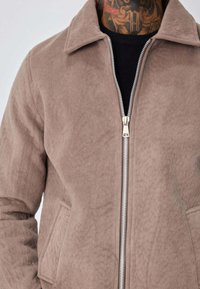 Veste marron en suède synthétique avec fermeture éclair sur le devant, poches latérales et col à revers. Surface texturée avec finition douce et mate.