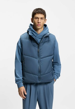 Junger Mann, der eine blaue Steppweste über einem blauen Kapuzenpullover und passende blaue Nadelstreifenhosen trägt, vor weißem Hintergrund stehend.