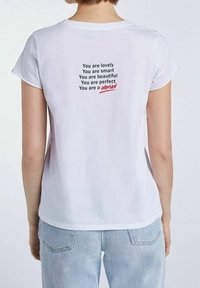 Camiseta de algodón blanca con cuello redondo, mangas cortas y afirmaciones impresas en negro y rojo en la parte posterior. Ajuste casual.