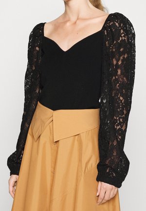 Czarny top z dekoltem w kształcie serca i przezroczystymi koronkowymi długimi rękawami, zestawiony z wysokim stanem spódnicy w kolorze karmelowym z asymetrycznym paskiem w talii.