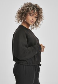 Felpa nera con scollatura rotonda e vita elastica, con spalle scese e un taglio rilassato. Indossata con pantaloni coordinati.