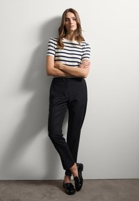Maglioncino a righe bianco e blu navy, pantaloni neri su misura e mocassini neri lucidi. Il modello posa con le braccia incrociate contro un muro.