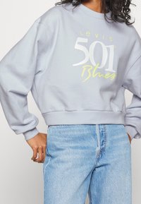 Personne portant un sweat-shirt Levi's 501 Blues violet clair et un jean taille haute bleu clair, posant les mains près des hanches.