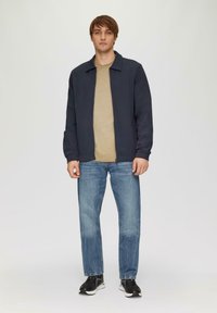 Veste zippée marine sur une chemise beige, associée à un jean bleu clair et des baskets noires. Tissu lisse avec une coupe décontractée et des détails minimalistes.