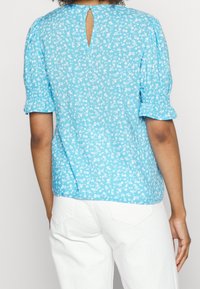 Blusa floreale blu con maniche a sbuffo e dettaglio a chiave sul retro, realizzata in tessuto leggero, con una texture morbida e un motivo floreale bianco.