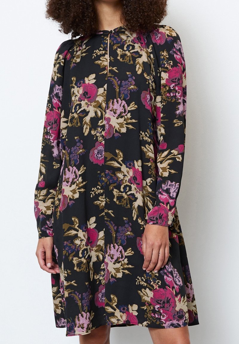 Personne portant une robe à manches longues, longueur genou, avec un fond noir et un imprimé floral audacieux rose, violet et beige, debout contre un mur uni.