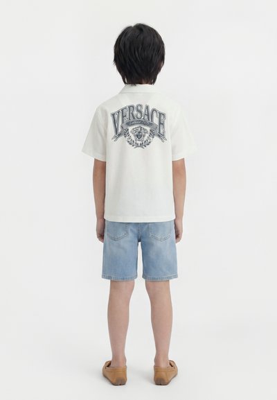 Παιδί που στέκεται με την πλάτη, φοράει λευκό πουκάμισο Versace με λογότυπο και έμβλημα λιονταριού, ανοιχτό μπλε τζιν σορτς και καφέ παπούτσια τύπου slip-on.