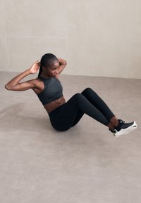 Top atlético negro y mallas, tejido texturizado, individuo realizando un abdominal sobre un suelo de color claro, vistiendo zapatillas atléticas negras.