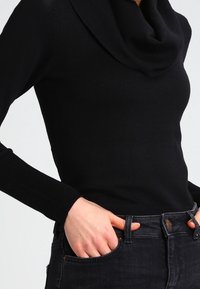 Pull noir avec un col bénitier, manches longues ajustées et poignets côtelés. Porté avec un jean foncé, mettant en valeur la texture lisse du tricot.
