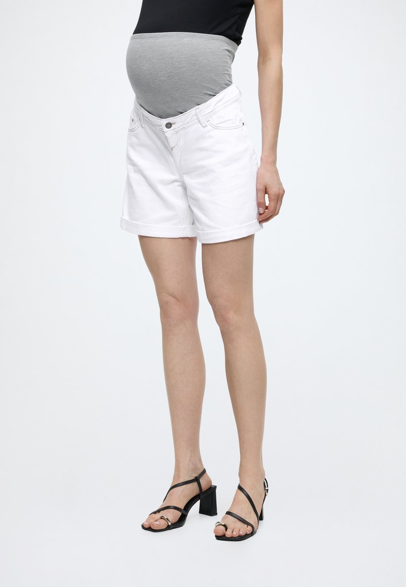 Έγκυος που φοράει λευκό σορτς εγκυμοσύνης με γκρι ελαστική μέση, μαύρο τοπ και μαύρα πέδιλα με τακούνι, στέκεται μπροστά σε άσπρο φόντο.