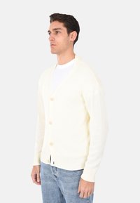 Cardigan color crema realizzato in maglia testurizzata, con scollatura a V, cinque bottoni sulla parte anteriore e polsini a coste, abbinato a jeans azzurri chiari.