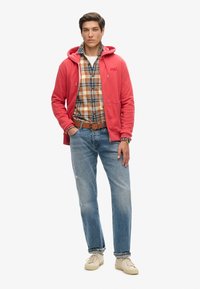 Koraalkleurige sweatshirt met capuchon, gedragen over een geruit overhemd, gecombineerd met lichtblauwe denim jeans en beige sneakers. Armband om de pols.