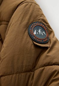 Manche de veste matelassée marron avec un patch rond noir et orange montrant des montagnes et les mots "Adventure Next Since 1982".