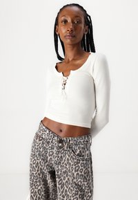Fehér bordás crop top, lekerekített nyakkivágással és fűzős elülső résszel, magas derekú leopárd mintás farmerral párosítva, ami klasszikus öt zsebes kialakítással rendelkezik.