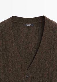 Bruine gebreide cardigan met een V-hals en een gestructureerd patroon. Voorzien van een enkele ronde zwarte knoop aan de voorkant en een label aan de binnenkant.