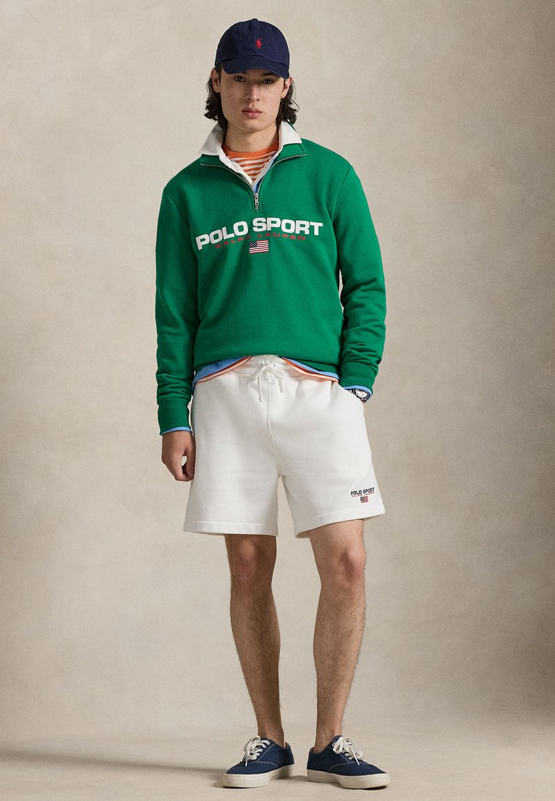 Green fleece sweatshirt with white zip collar featuring "POLO SPORT" print. White shorts and navy sneakers completed with a navy cap.