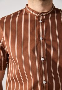Brown short-sleeve shirt with vertical white stripes, featuring a band collar and white buttons down the front. Smooth, woven fabric texture.