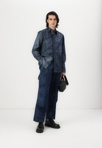 Camicia blu scuro a fantasia con maniche lunghe e tasche, abbinata a pantaloni larghi dello stesso tono. Borsa accessorio nera e scarpe.