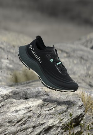 Sort Columbia trail-løbesko med turkis snørebånd svævende over klippefyldt terræn med små planter og sløret baggrund.