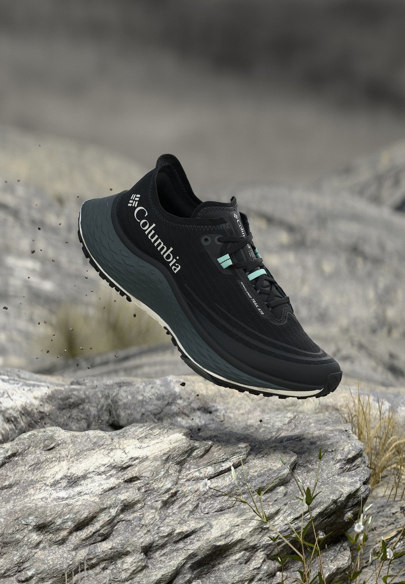 Μαύρο παπούτσι για τρέξιμο στο μονοπάτι Columbia με βεραμάν κορδόνια να αιωρείται πάνω από πέτρινο έδαφος με μικρά φυτά και θαμπό φόντο.